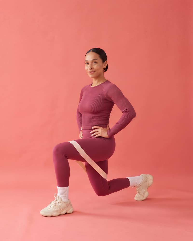 A smiling woman in a berry top and leggings holds a lunge, a mini band above her knees, in a coral photo studio.